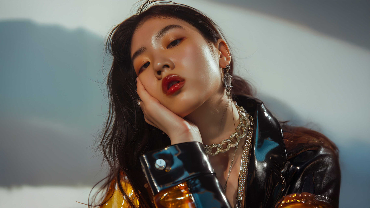 A young woman with long dark hair rests her face on her hand, gazing confidently at the camera. She wears bold makeup, layered necklaces, hoop earrings, and a shiny black and yellow jacket. Soft light casts shadows behind her.