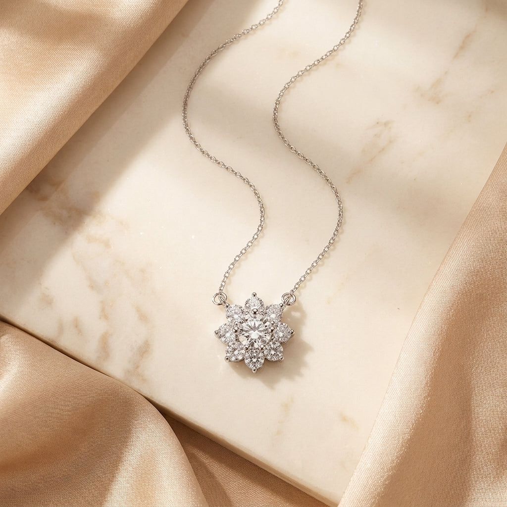 A delicate silver chain necklace with a snowflake-shaped pendant set with clear stones, displayed on a cream marble slab with draped champagne silk fabric.